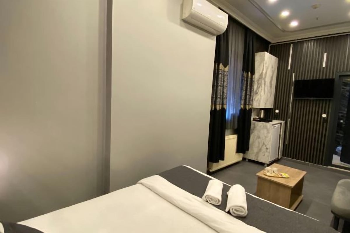 A hotel room with a bed, a small wooden side table, a chair, a desk with a lamp, and a TV mounted on the wall.