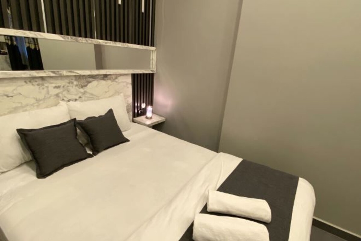 A neatly made bed with white and dark gray bedding, a bedside table with a lamp, and a large mirror above the bed.