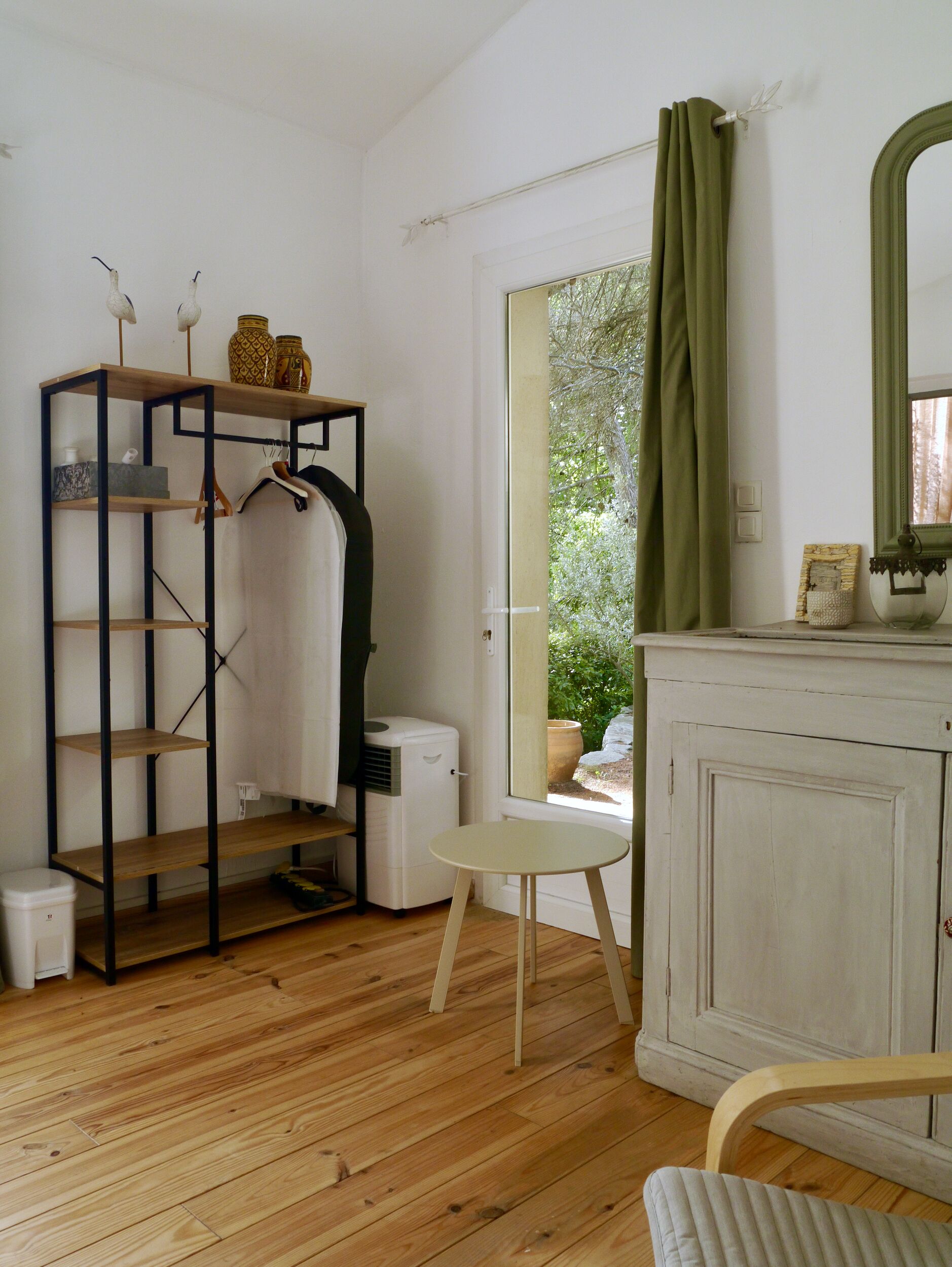 A room with a wooden floor, a white wardrobe, a small table, a hanging rack with clothes, and a window with curtains.