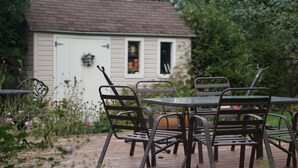 Un patio con una mesa y sillas, una caseta pequeña con puerta y ventanas, y un jardín con diversas plantas y flores.