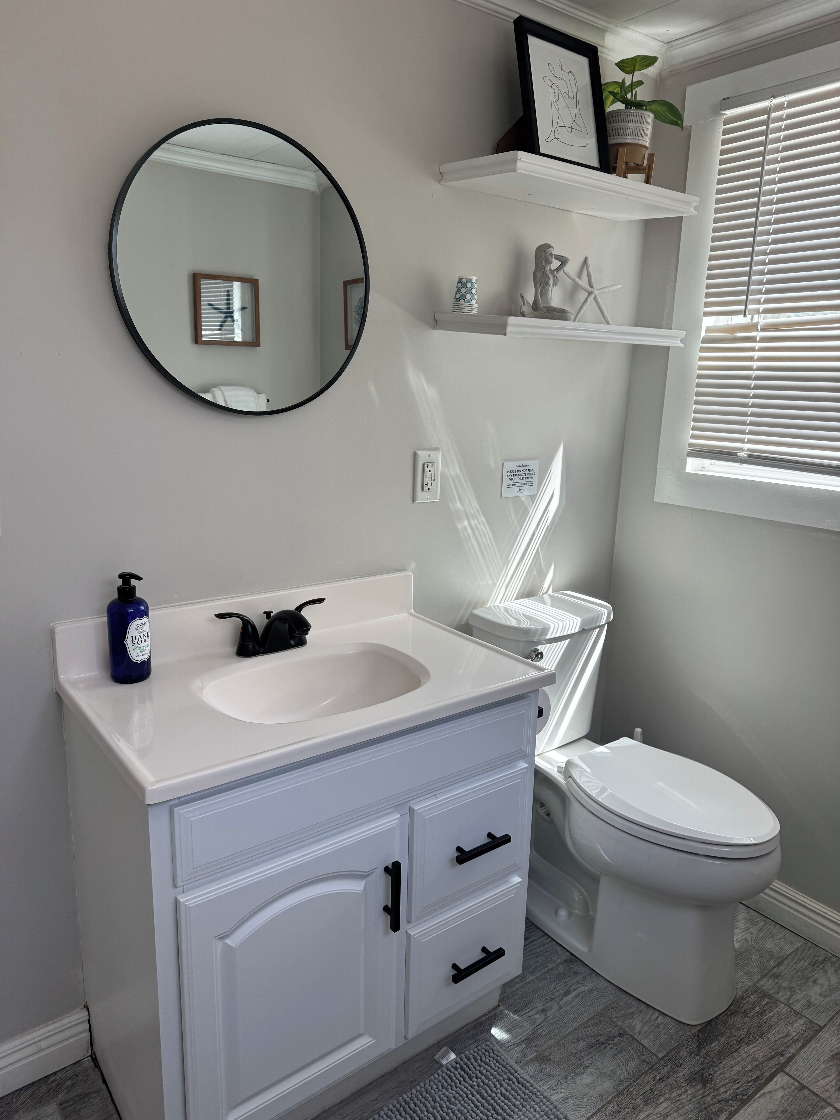 A modern bathroom with a white vanity, a round mirror, a toilet, and a window with blinds.