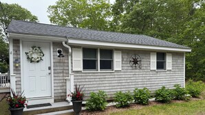A single-story house with a white door, a front porch, and a small garden area with potted plants. - Cozy Cottage walk to Menauhant Beach (East Falmouth)
