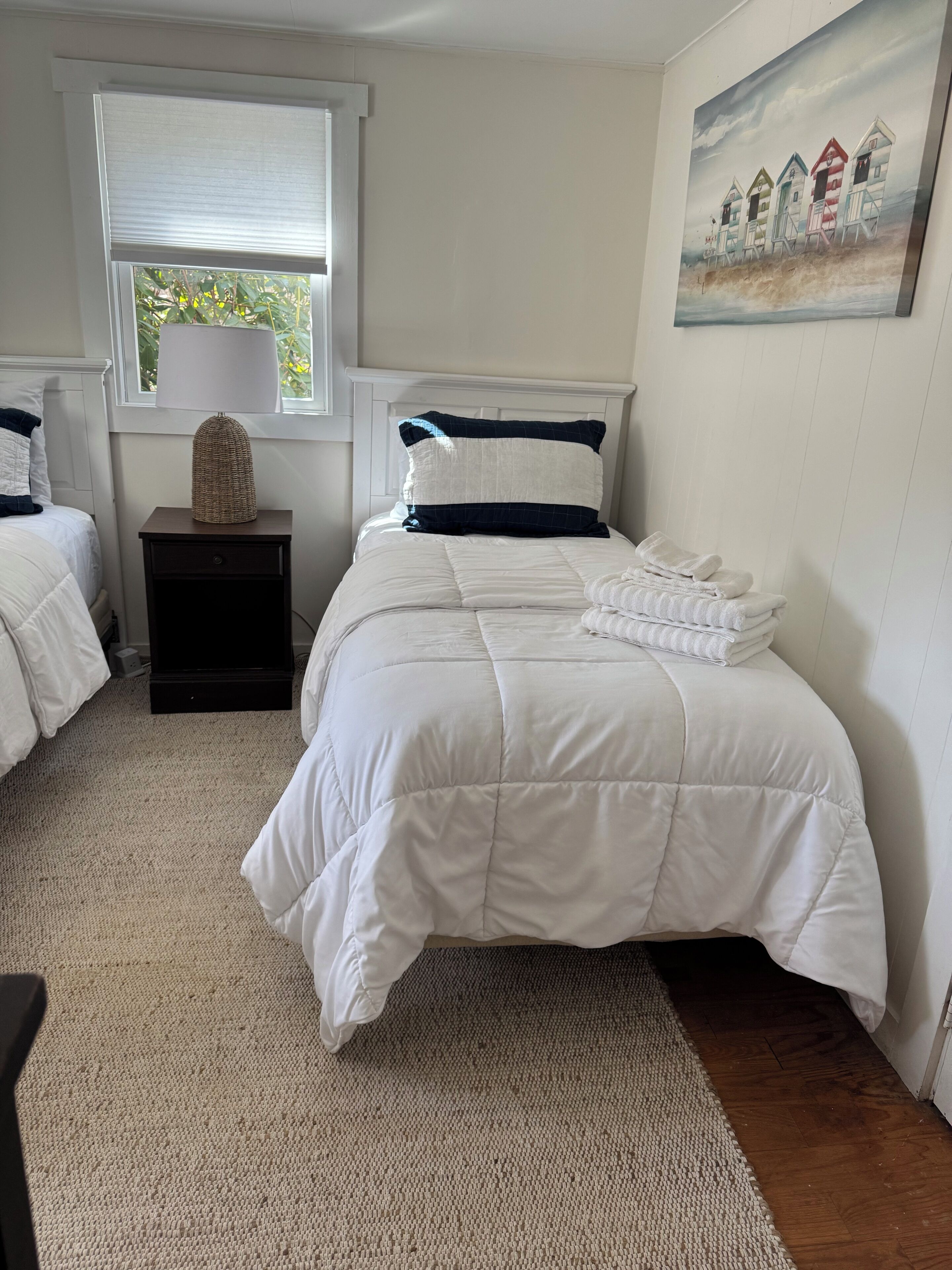 A bedroom with two beds, a window with blinds, a nightstand with a lamp, and a painting on the wall.
