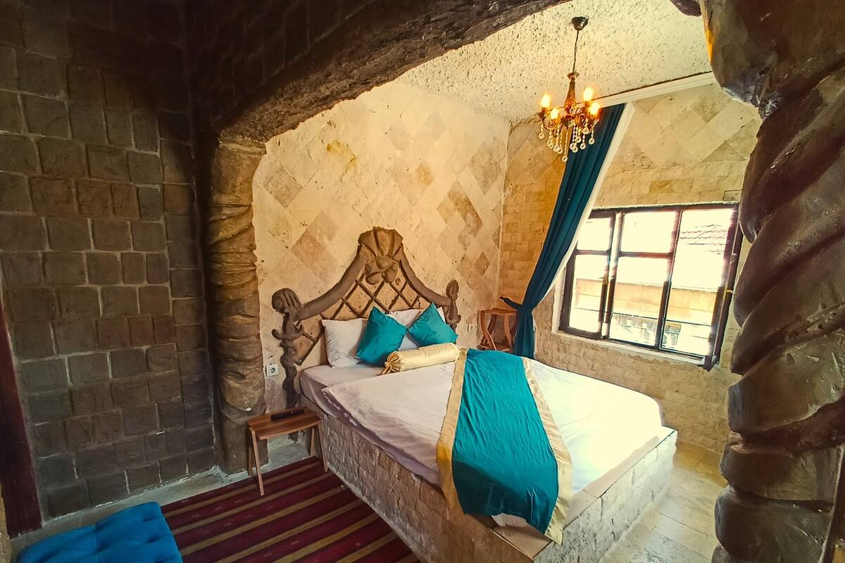 Une chambre avec un mur en pierre, un lit en bois, un lustre et une vue sur l’extérieur par une fenêtre.