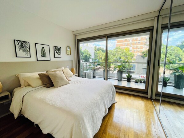 A bedroom with a large bed, a nightstand, and a window with a view of buildings and trees.