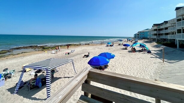 A beach with a wooden boardwalk, beach umbrellas, and a building with balconies. - 2nd Row, Private Pool and Jacuzzi! (Kure Beach)