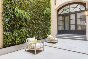 A courtyard with a lush green wall, two white wicker chairs, and a large arched window with curtains.