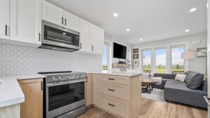 A modern kitchen with white cabinets, a built-in microwave, and a stainless steel oven. There is a grey sofa and a TV in the adjacent living area.