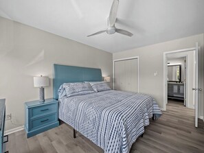 A bedroom with a bed, nightstand, lamp, ceiling fan, and a door leading to a bathroom.
