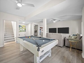 A modern living room with a pool table, a sofa, a glass coffee table, and a television. - Sea Glass (B) Upscale Beachfront Retreat & Pool - 5BR (Emerald Isle)