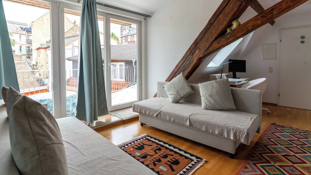 A modern living room with a sofa, a patterned rug, and a view of the outside through large windows. - Attic Apartment at Schlosspark (Wiesbaden)