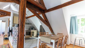 A dining area with a wooden table and chairs, a kitchen in the background, and a sloped ceiling with exposed beams. - Attic Apartment at Schlosspark (Wiesbaden)