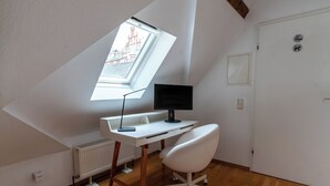 A room with a sloped ceiling, a white desk with a computer, a white chair, and a window with a view of buildings. - Attic Apartment at Schlosspark (Wiesbaden)
