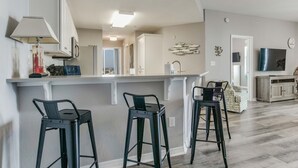 A modern kitchen with a bar area, two bar stools, a refrigerator, and a television. - Bayfront Bliss: Top-Floor Condo with Stunning Views (Gulf Shores)