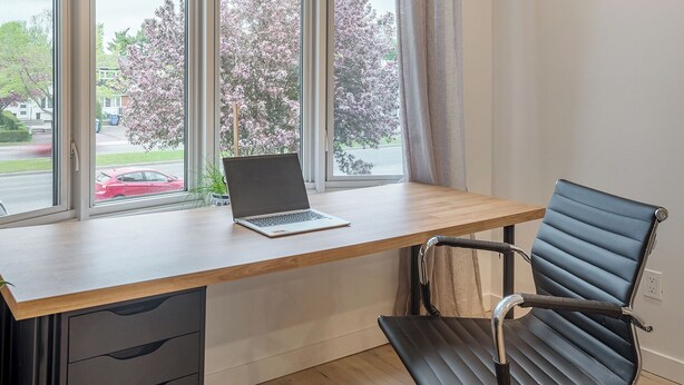 Un espacio de trabajo moderno con un ordenador portátil sobre un escritorio de madera, una silla de oficina negra y grandes ventanales que ofrecen vistas a una calle y flores de cerezo.