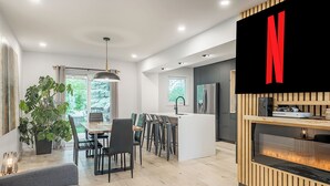 Una cocina moderna con una isla integrada, un comedor con una mesa grande y sillas, y una sala de estar con sofá y televisor montado en la pared.