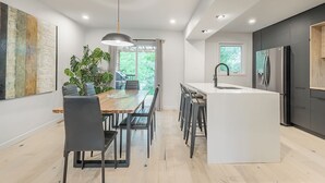 A modern kitchen with a central island, a dining area with chairs, a large window, and a plant.