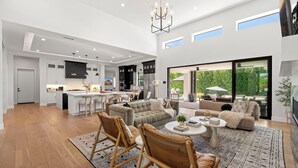 Een ruime, moderne keuken met een centraal eiland, een eetgedeelte met een kroonluchter, en een woonkamer met een groot raam dat uitkijkt op groen.