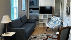 A living room with a sofa, a chair, a TV, a lamp, and a bed in the background. - Walk To Goodspeed Opera House! (East Haddam)
