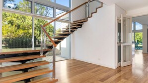 Un escalier moderne avec des rampes en verre et des marches en bois, menant à une pièce dotée de grandes fenêtres et offrant une vue sur les arbres.