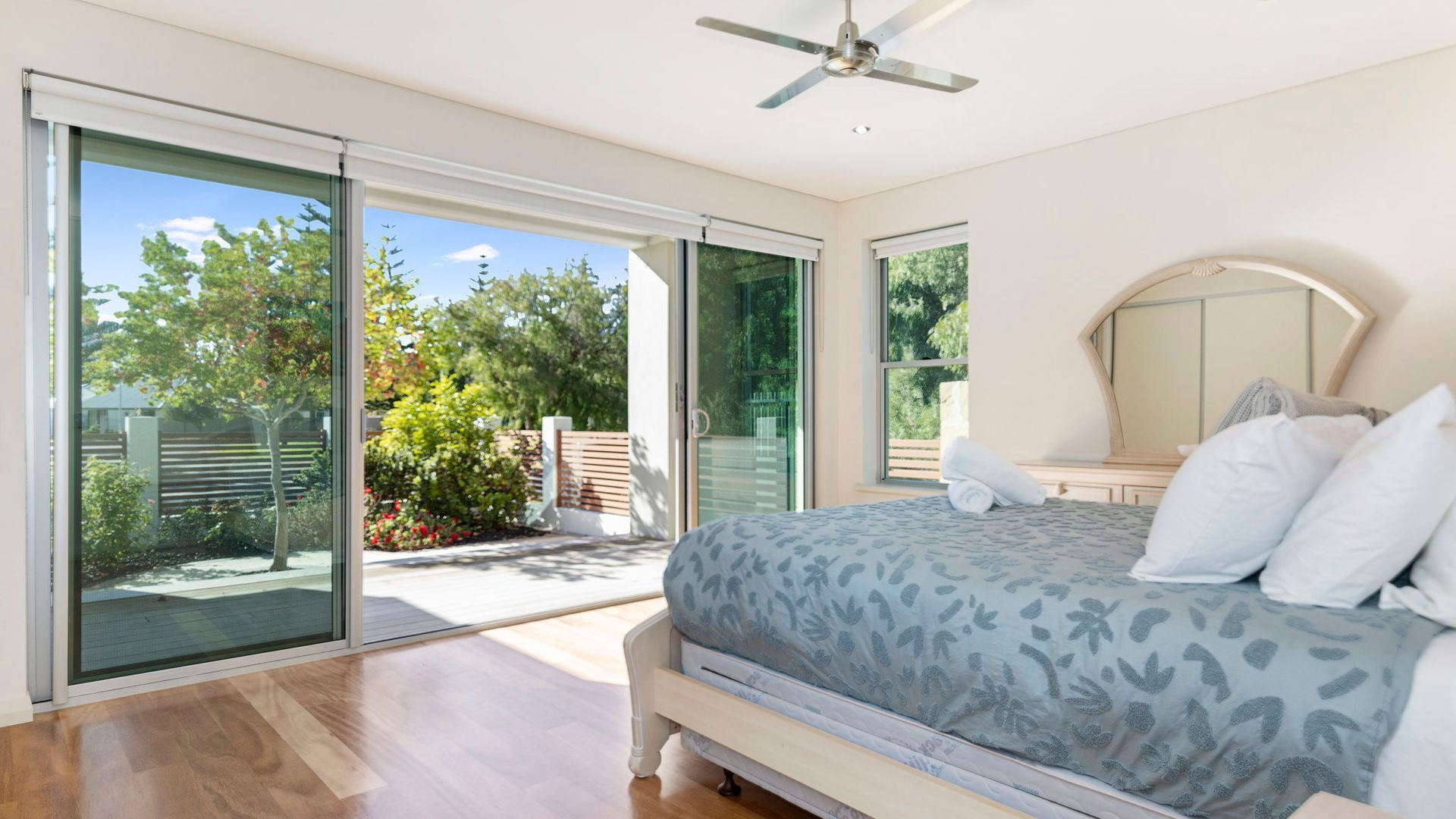 A bedroom with a bed, a ceiling fan, and a sliding glass door leading to a balcony with a view of trees and a fence.