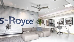 Een modern interieur met een groot S-Royce logo op de muur, een grijze modulaire bank, een plafondventilator en afbeeldingen van luchtvaartthema in lijsten.