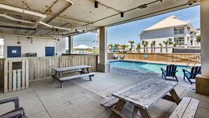 An outdoor pool area with wooden picnic tables and lounge chairs, a covered patio with a pool, and a view of a residential building.