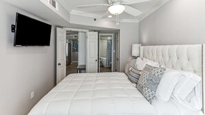 A bedroom with a large bed, a ceiling fan, a TV mounted on the wall, and a bathroom visible through an open door.