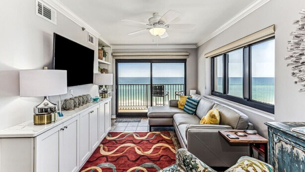 A modern living room with a large TV, a ceiling fan, a sofa with cushions, a red patterned rug, and a view of the ocean through a balcony door.