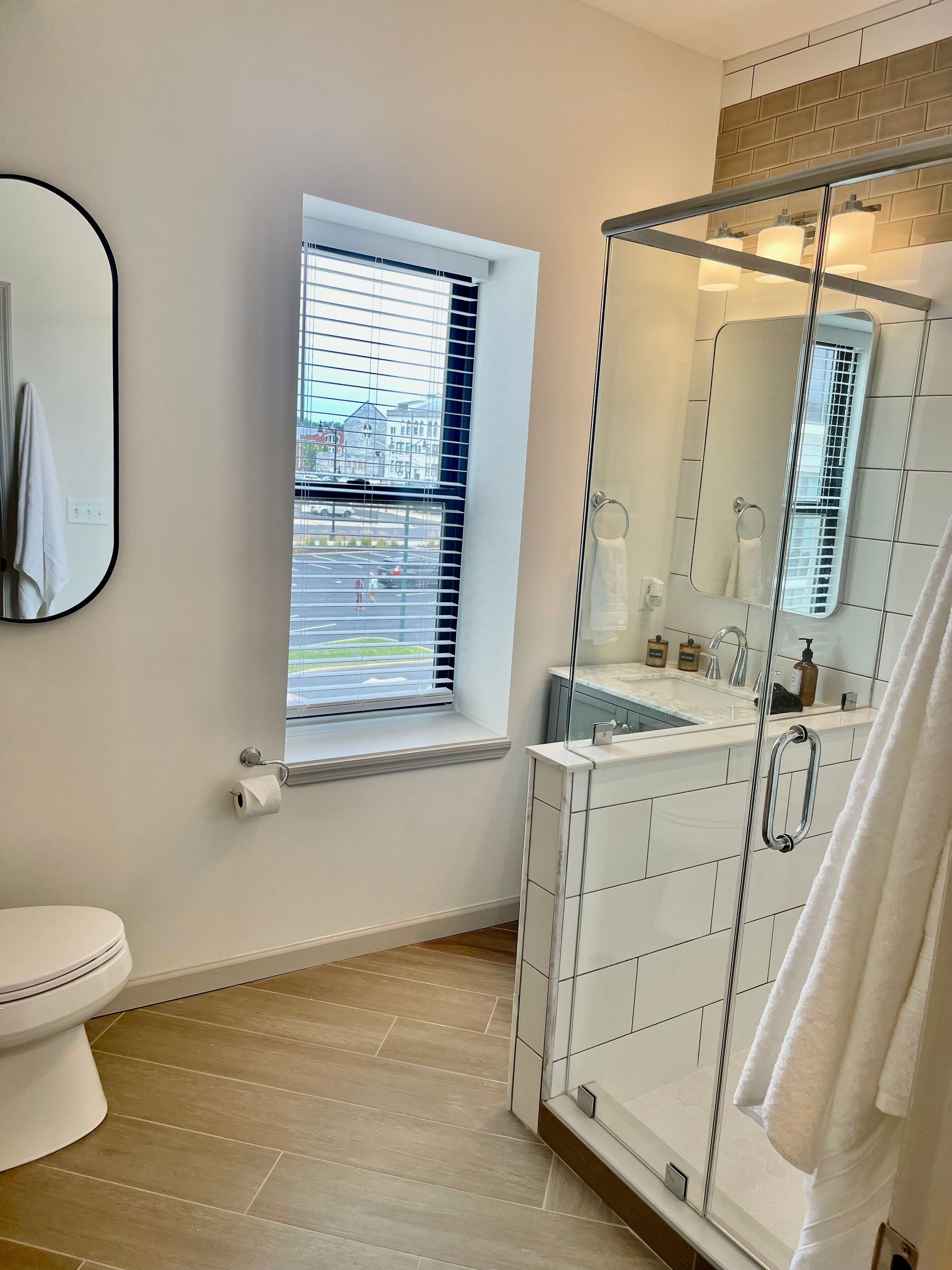 Un baño moderno con inodoro, ventana con persianas, cabina de ducha de vidrio y espejo.