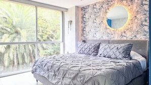 Ett modernt sovrum med ett stort fönster, en säng med ett geometriskt mönstrat överkast, en rund väggspegel och en väggdesign som liknar marmor.
