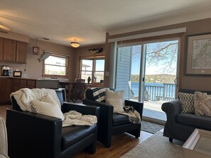 A cozy living room with two black leather sofas, a wooden dining table, and a kitchen area with wooden cabinets. A large sliding glass door leads to a deck with a view of a lake.