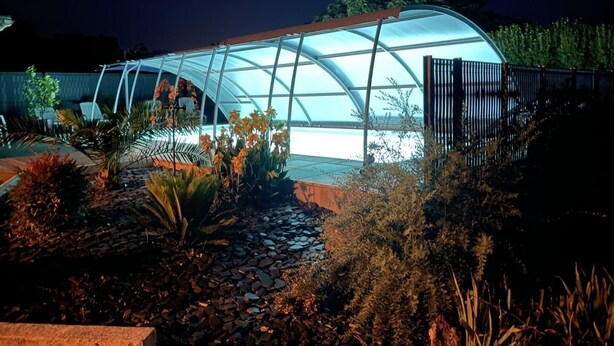 Una estructura de invernadero con techo transparente, rodeada por un jardín ajardinado con diversas plantas y un sendero.