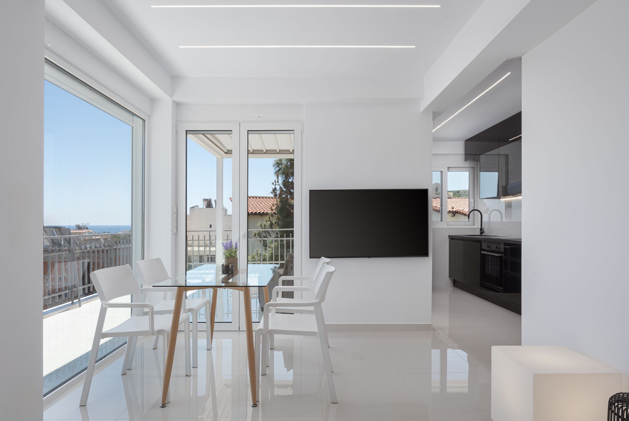 A modern living room with a glass table and white chairs, a large TV, and a kitchen area with a black countertop.