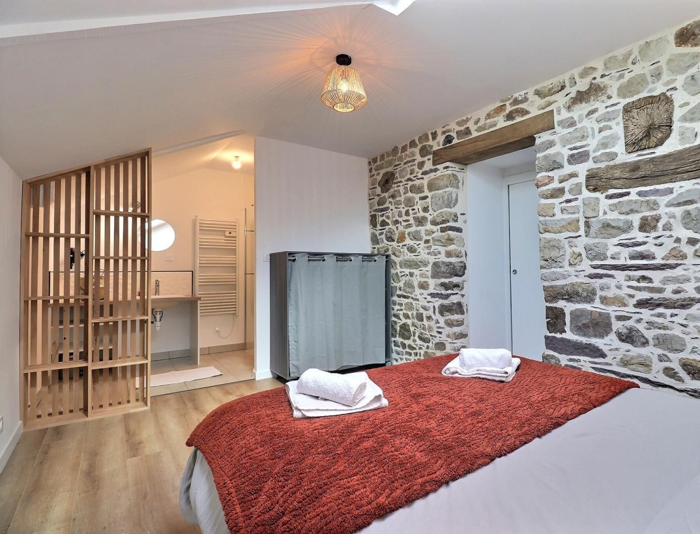 A bedroom with a stone wall, a wooden wardrobe, a bed with a red blanket, and a towel on the bed.