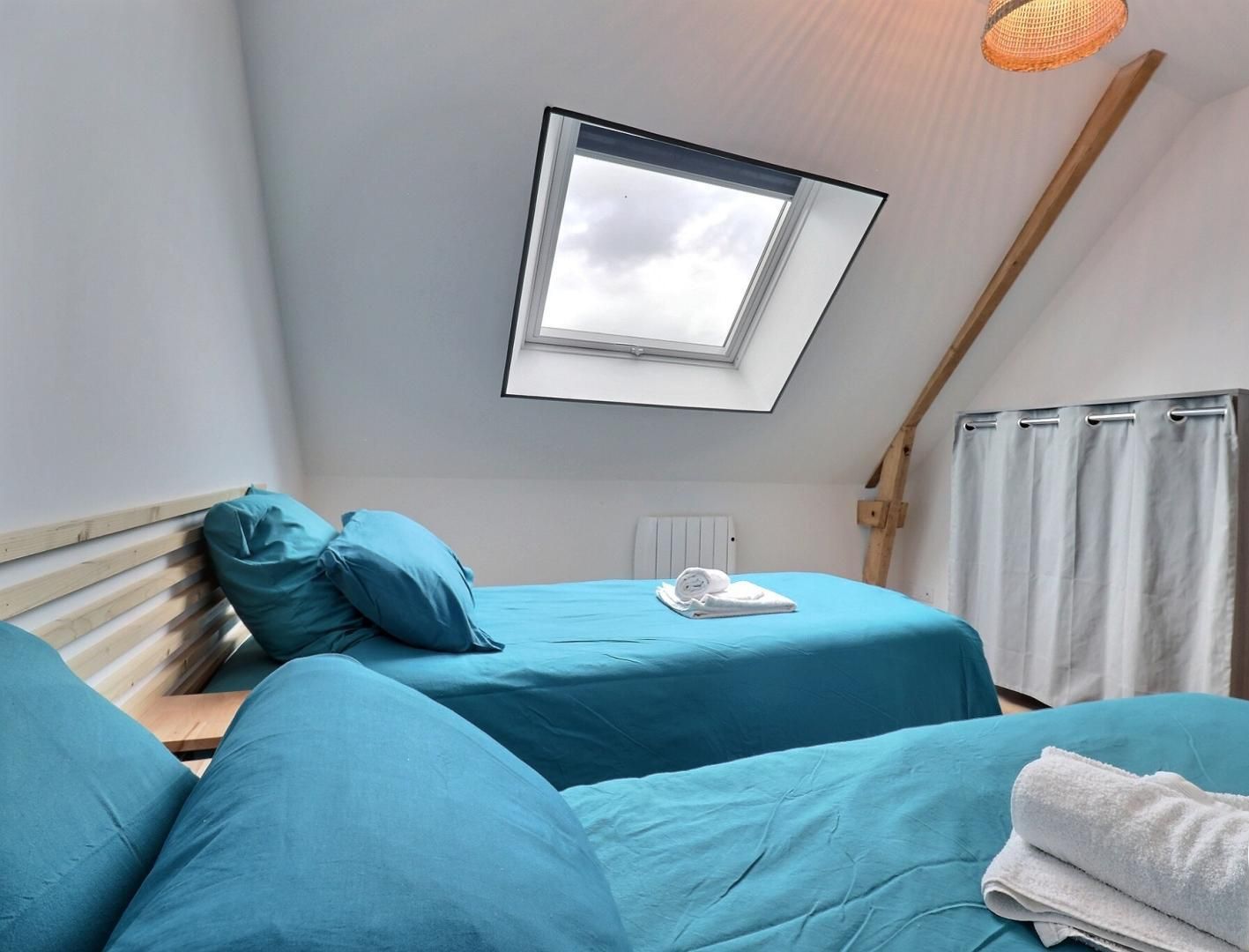 A bedroom with two beds, a skylight, a wooden ceiling beam, and a white curtain.