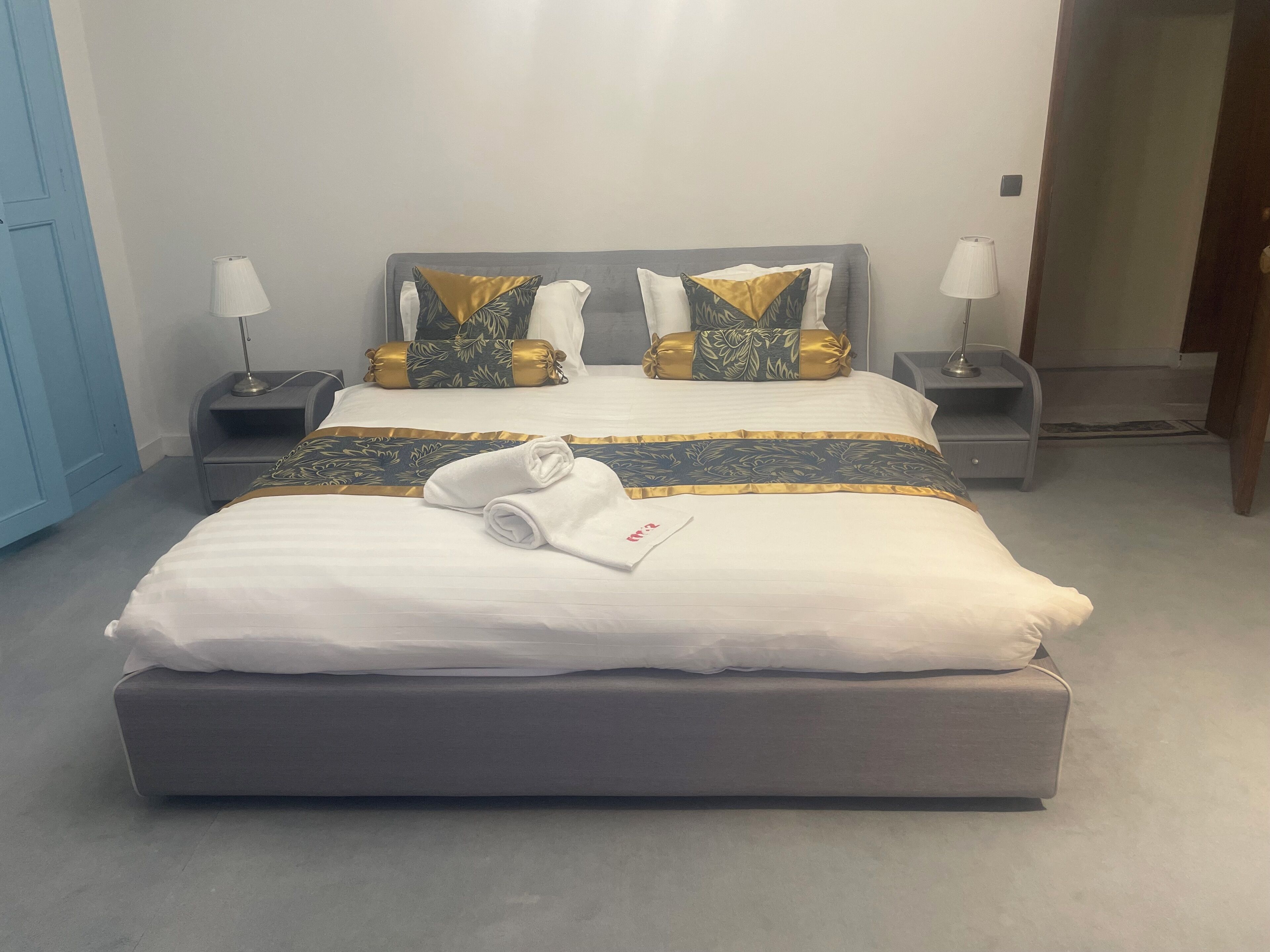 Una cama bien hecha con edredón blanco, dos almohadas con diseños dorados y verdes, y una toalla doblada sobre la cama. Hay dos mesitas de noche con lámparas, un cabecero gris y una puerta azul al fondo.