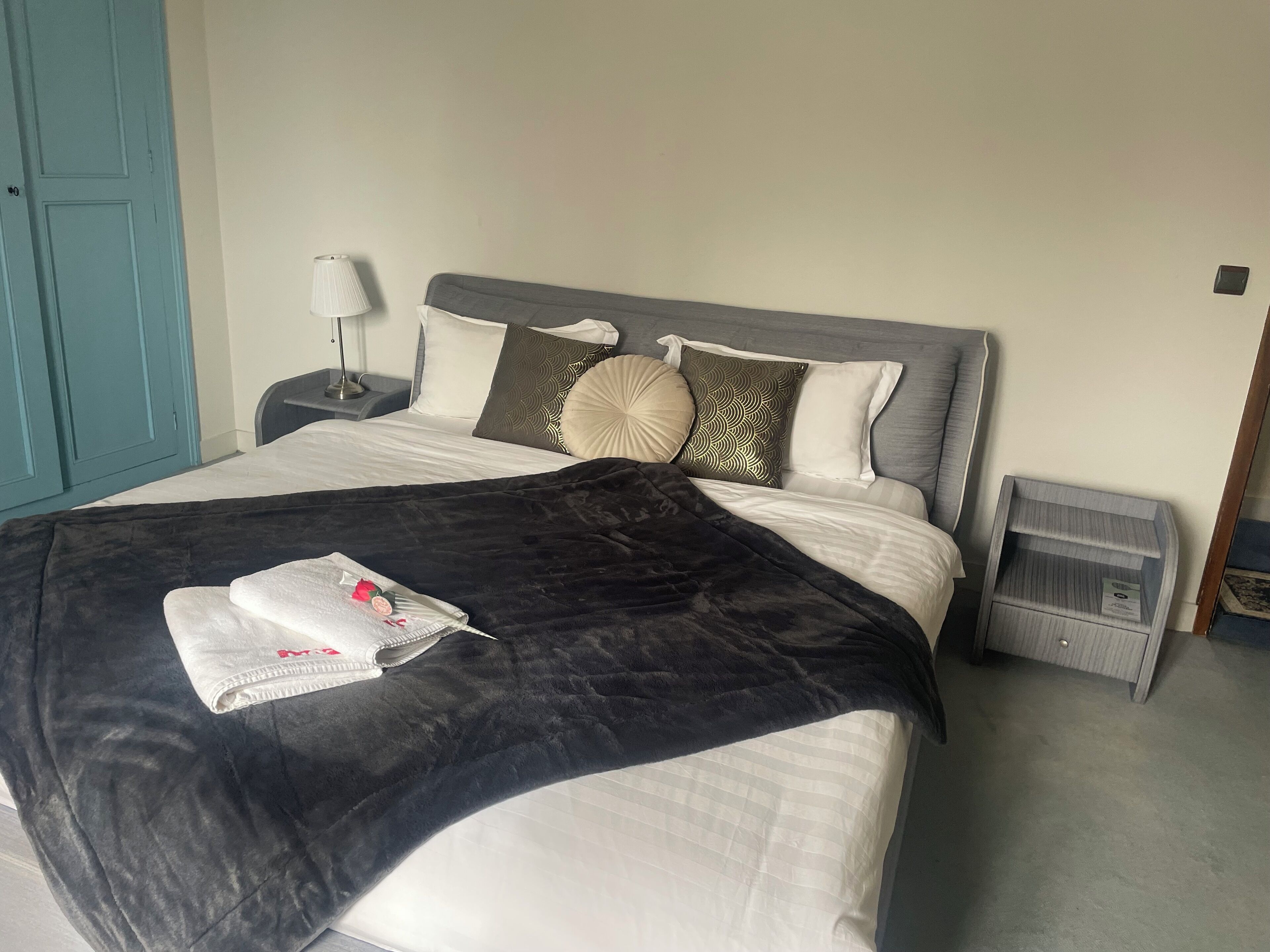 A neatly made bed with a black blanket and a white pillow with a red rose. There is a bedside table with a lamp and a small drawer.