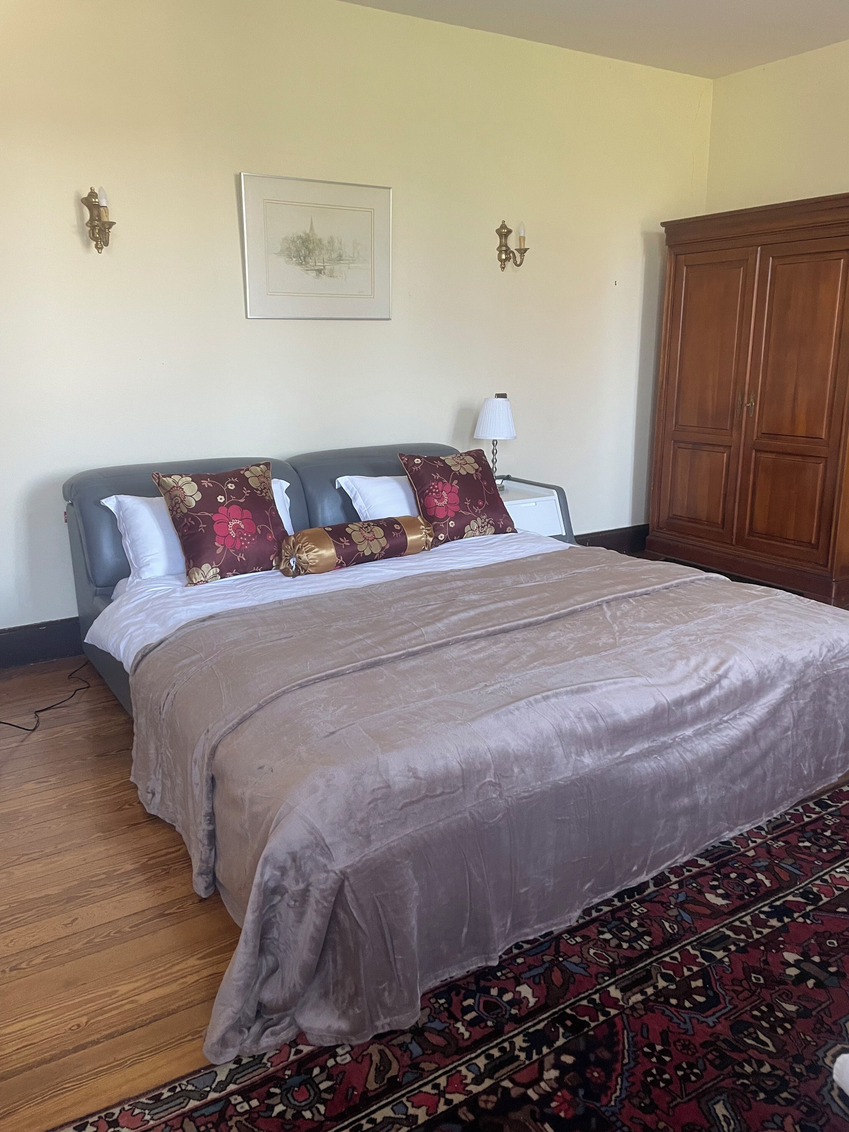 A bedroom with a large bed, a wooden wardrobe, a patterned rug, and a framed picture on the wall.