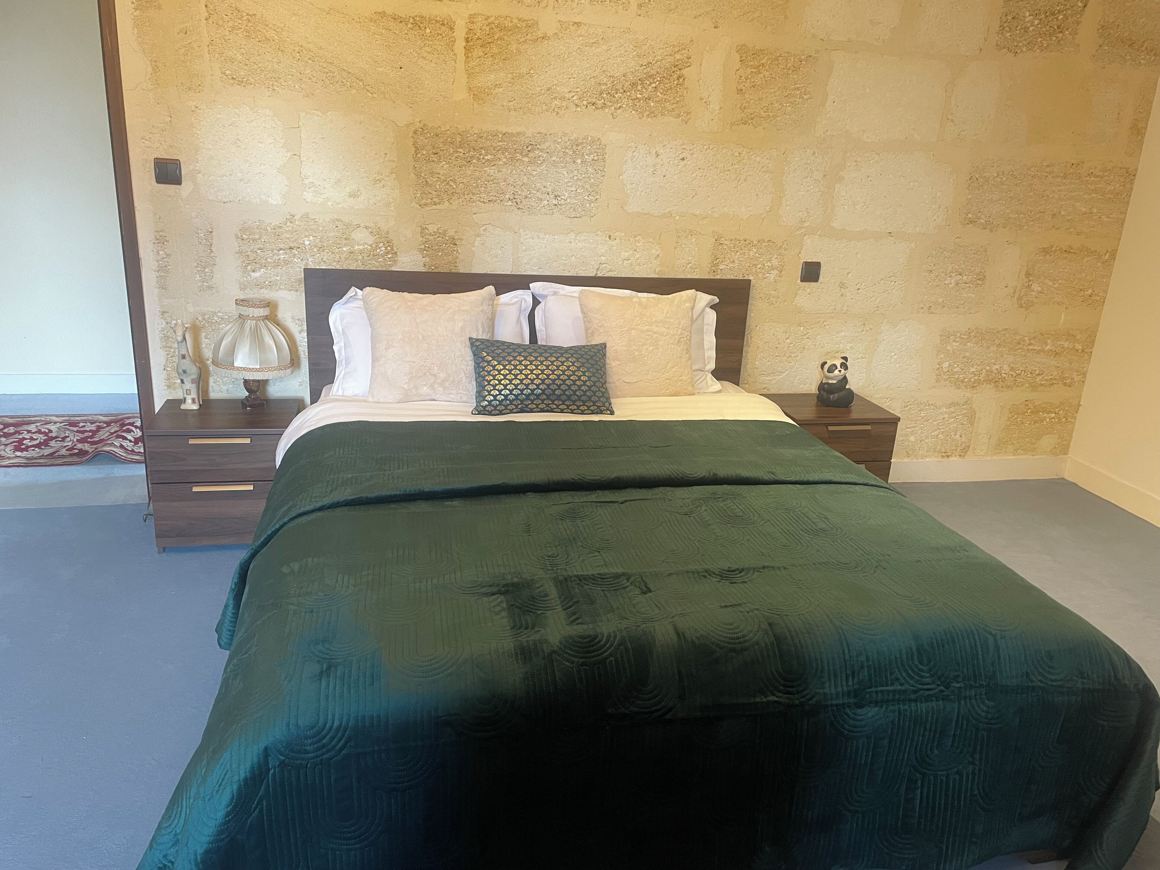A bedroom with a stone wall, a bed with a green bedspread, white pillows, and a small bedside table with a lamp and a panda figurine.