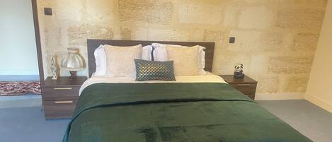 A bedroom with a stone wall, a bed with a green bedspread, white pillows, and a small bedside table with a lamp and a panda figurine.