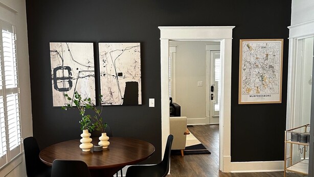Een moderne eetruimte met een donkere muur, houten vloer en een ronde tafel met stoelen. Er hangen twee abstracte schilderijen en een kaart aan de muur.