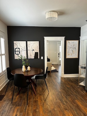 A modern dining area with a dark wall, wooden flooring, and a round table with chairs. There are two abstract paintings and a map on the wall.