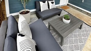Een overdekt terras met een donkerblauwe muur, een plant in pot, een grijze bank met decoratieve kussens en een bijpassend bijzettafeltje.