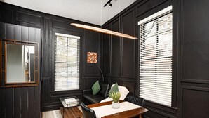 Una habitación con paredes revestidas de madera oscura, un sofá de cuero negro, una mesa de comedor de madera con sillas, un espejo y una ventana con persianas.