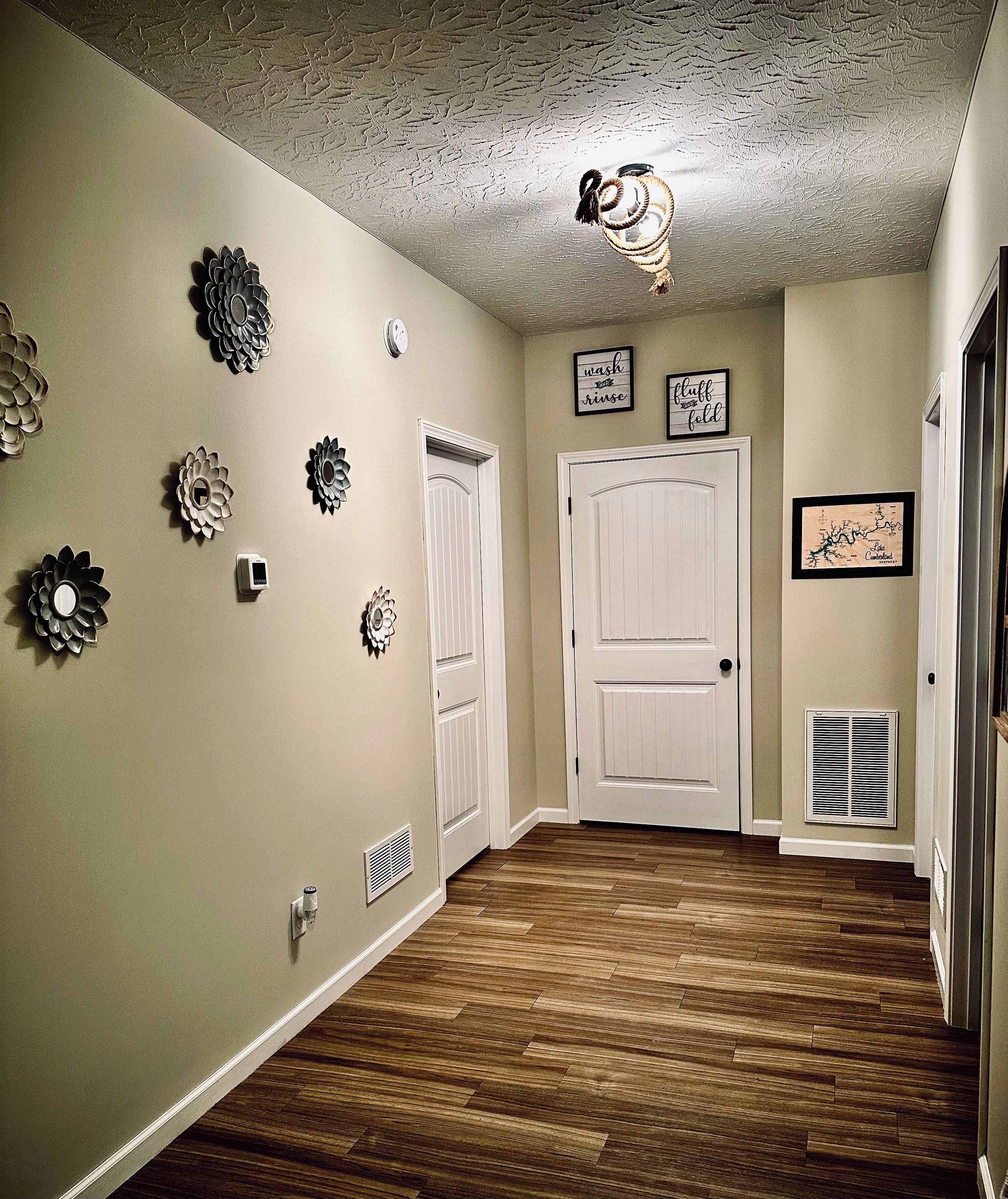 Un couloir doté d’un plancher en bois, de murs de couleur beige, d’œuvres d’art décoratives accrochées au mur, d’un ventilateur de plafond et d’une porte au-dessus de laquelle est fixée une enseigne.