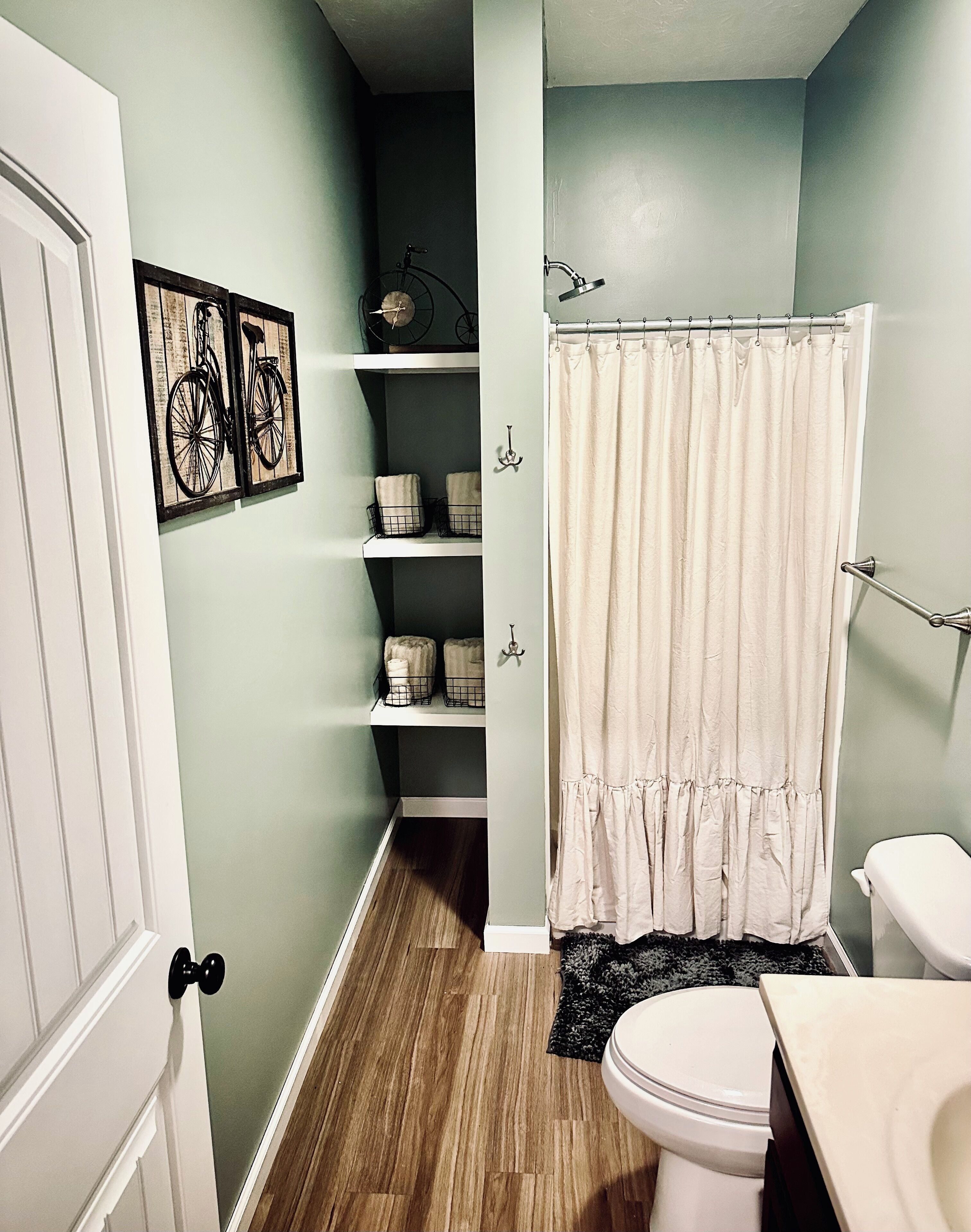 A narrow bathroom with a wooden floor, a white toilet, a white sink, a beige shower curtain, and shelves with towels and baskets.