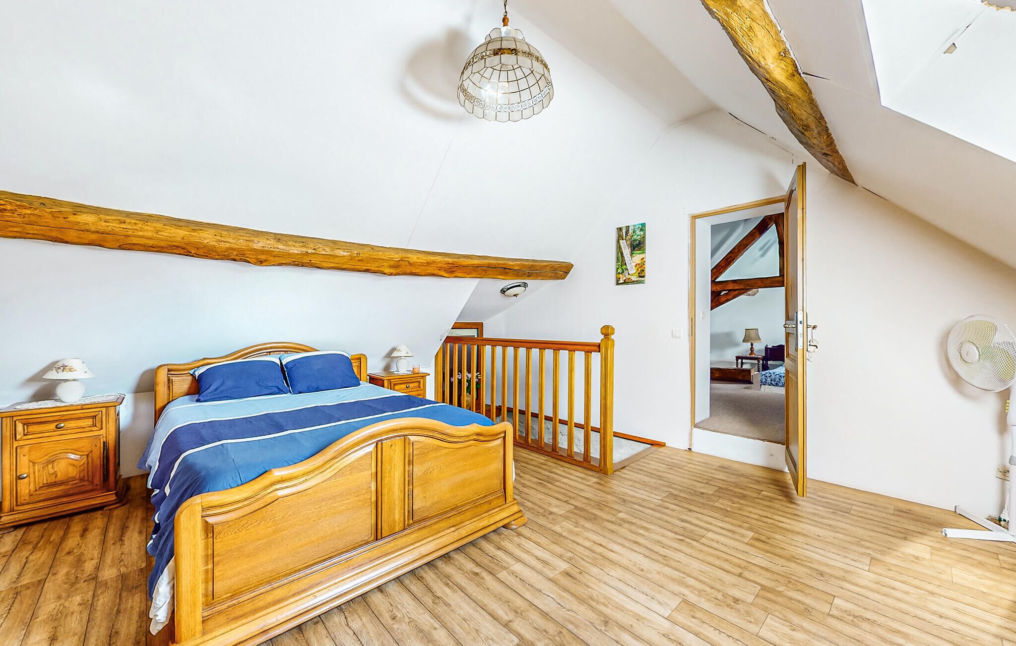 A bedroom with a wooden bed, a nightstand, a lamp, a wooden ceiling beam, and a door leading to another room.