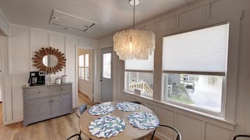 A dining area with a round table, four placemats, a chandelier, a mirror, and a cabinet with a coffee maker.
