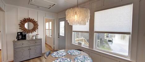 A dining area with a round table, four placemats, a chandelier, a mirror, and a cabinet with a coffee maker.
